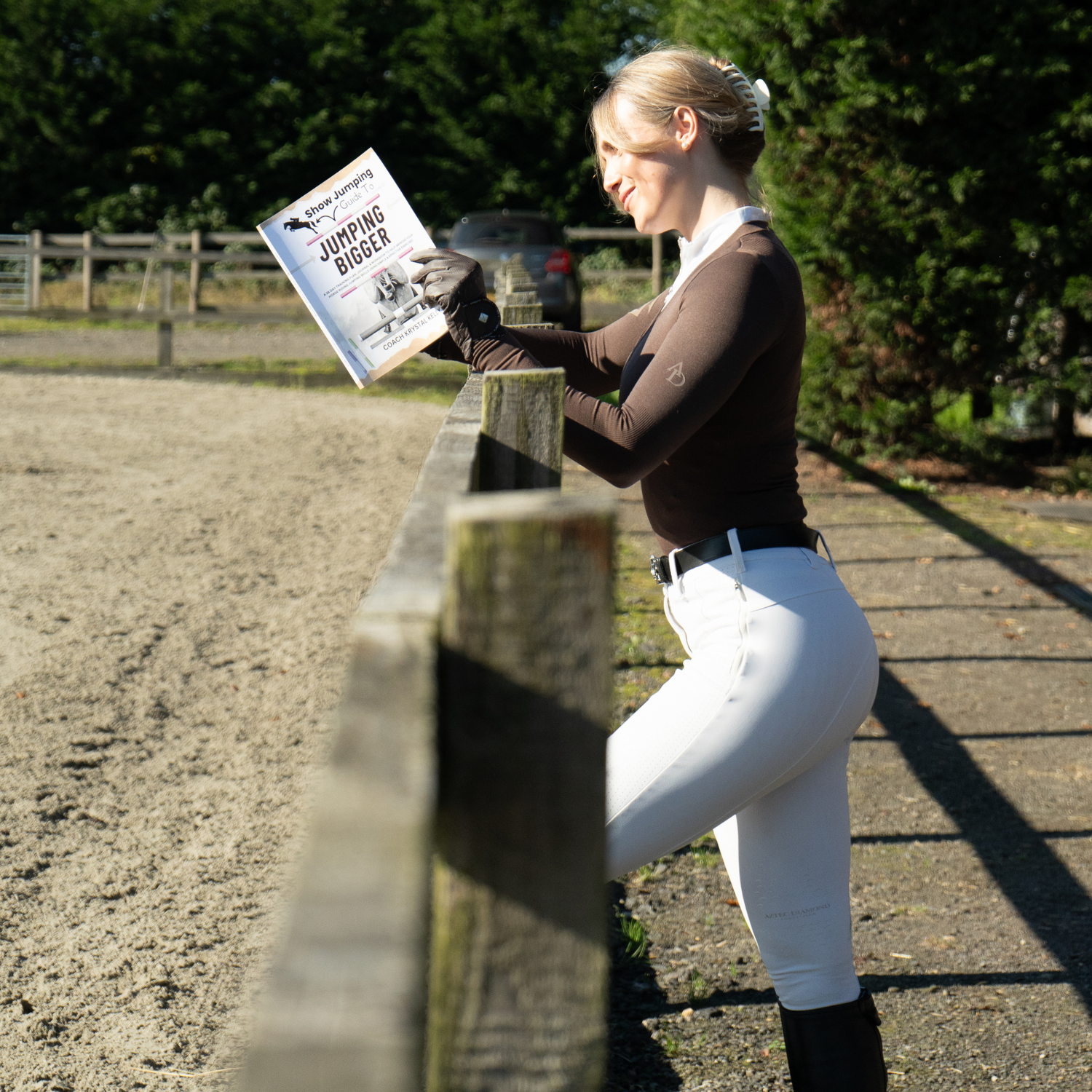 A woman in equestrian attire reads a book titled 'Jumping Bigger'