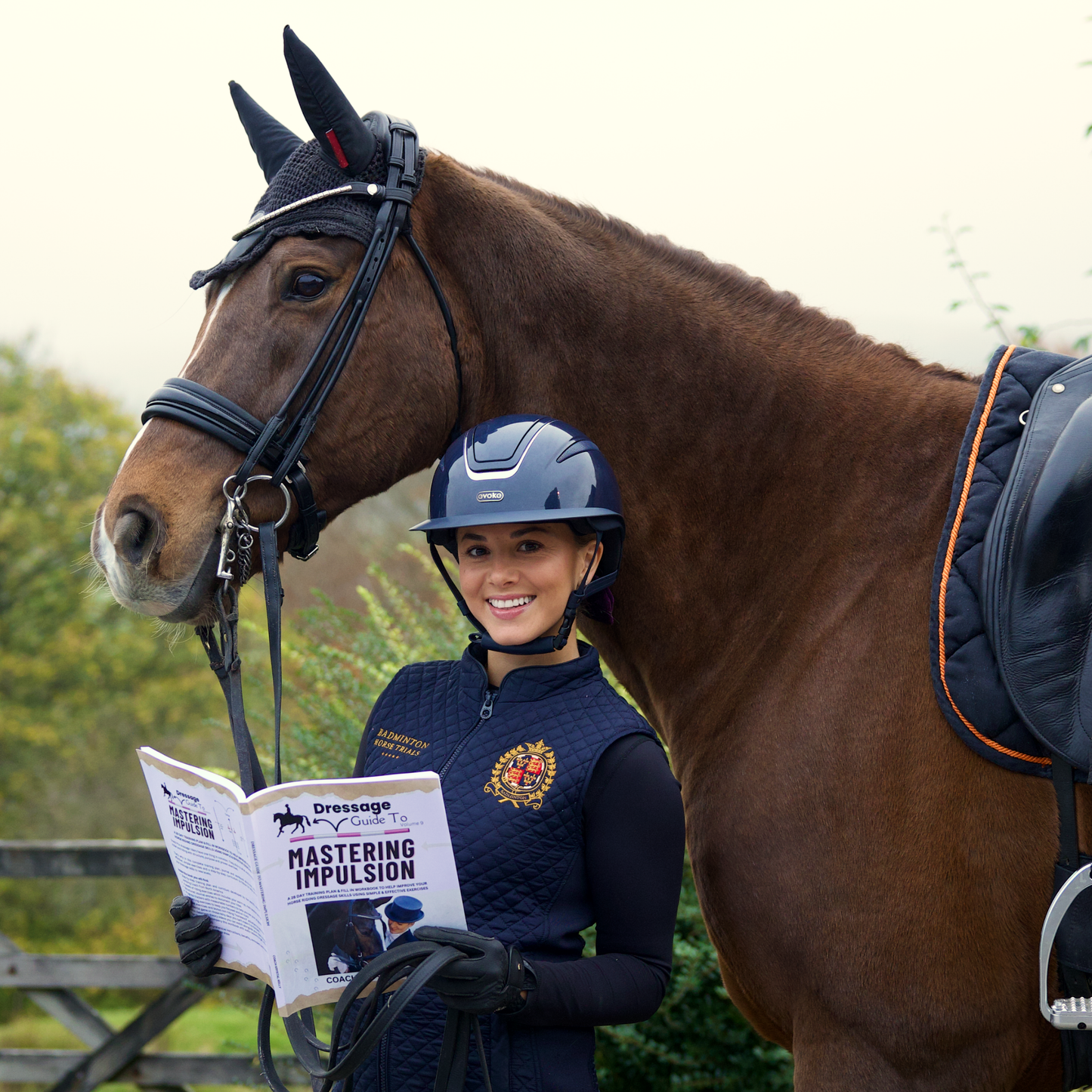 A woman in equestrian attire holding a book titled Mastering Impulsion